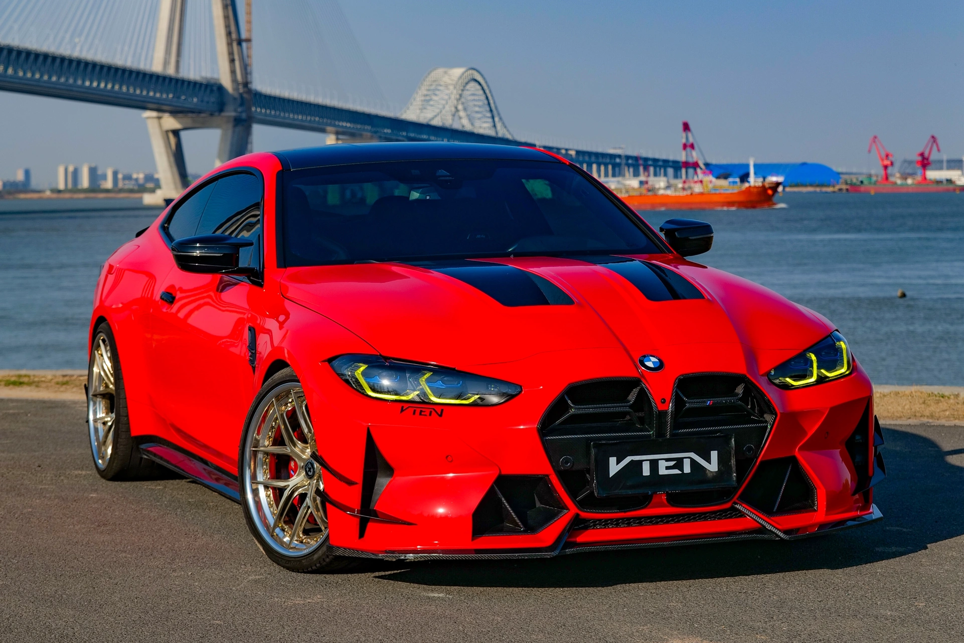 Red BMW M4 G82 sports car with 'VTEN' branding on a road by a bridge and water.