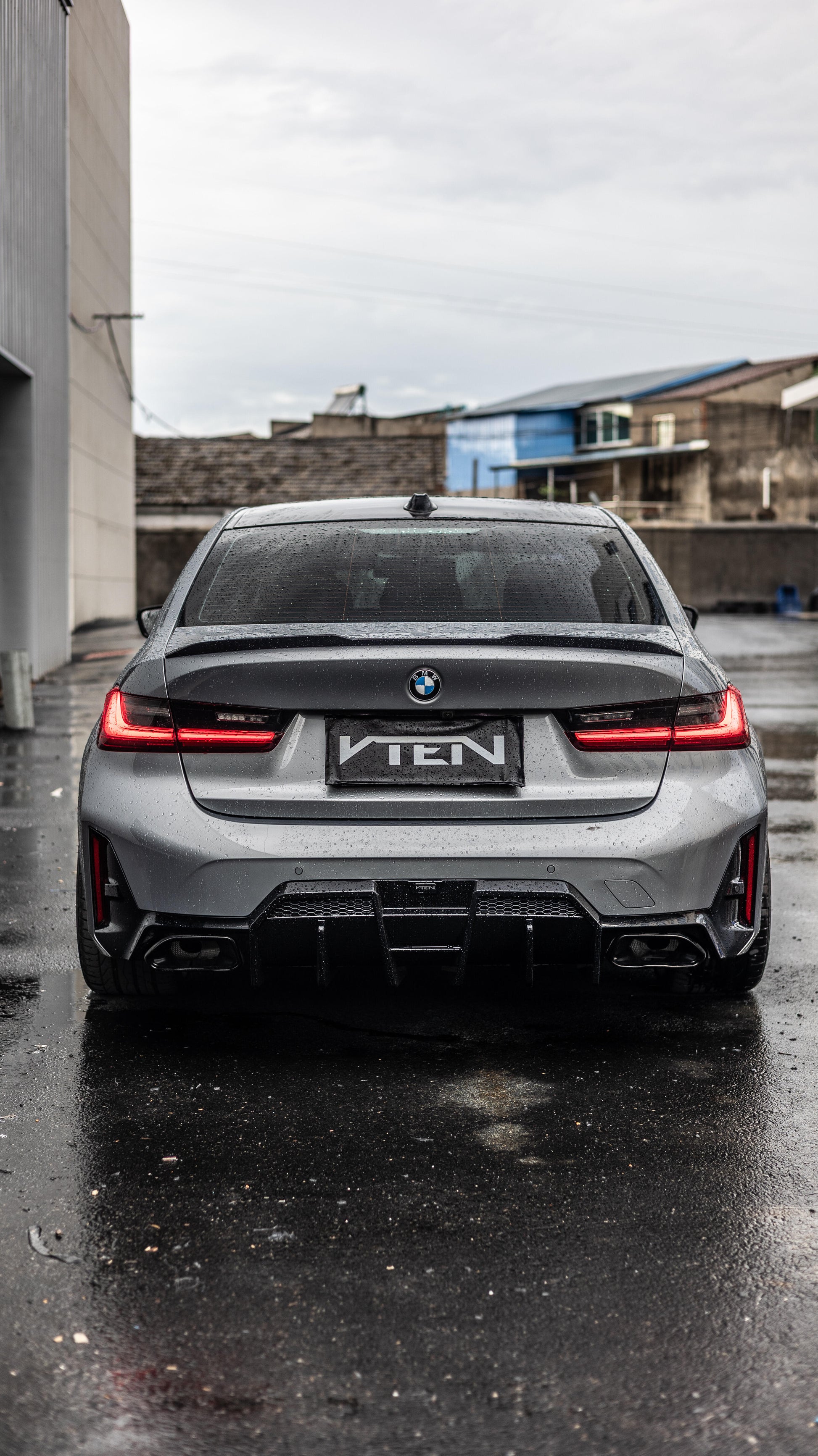 Rear view of a silver BMW 3 Series G20 LCI sedan parked outdoors on wet pavement, with a custom black diffuser and a central plate reading “VTEN.”.