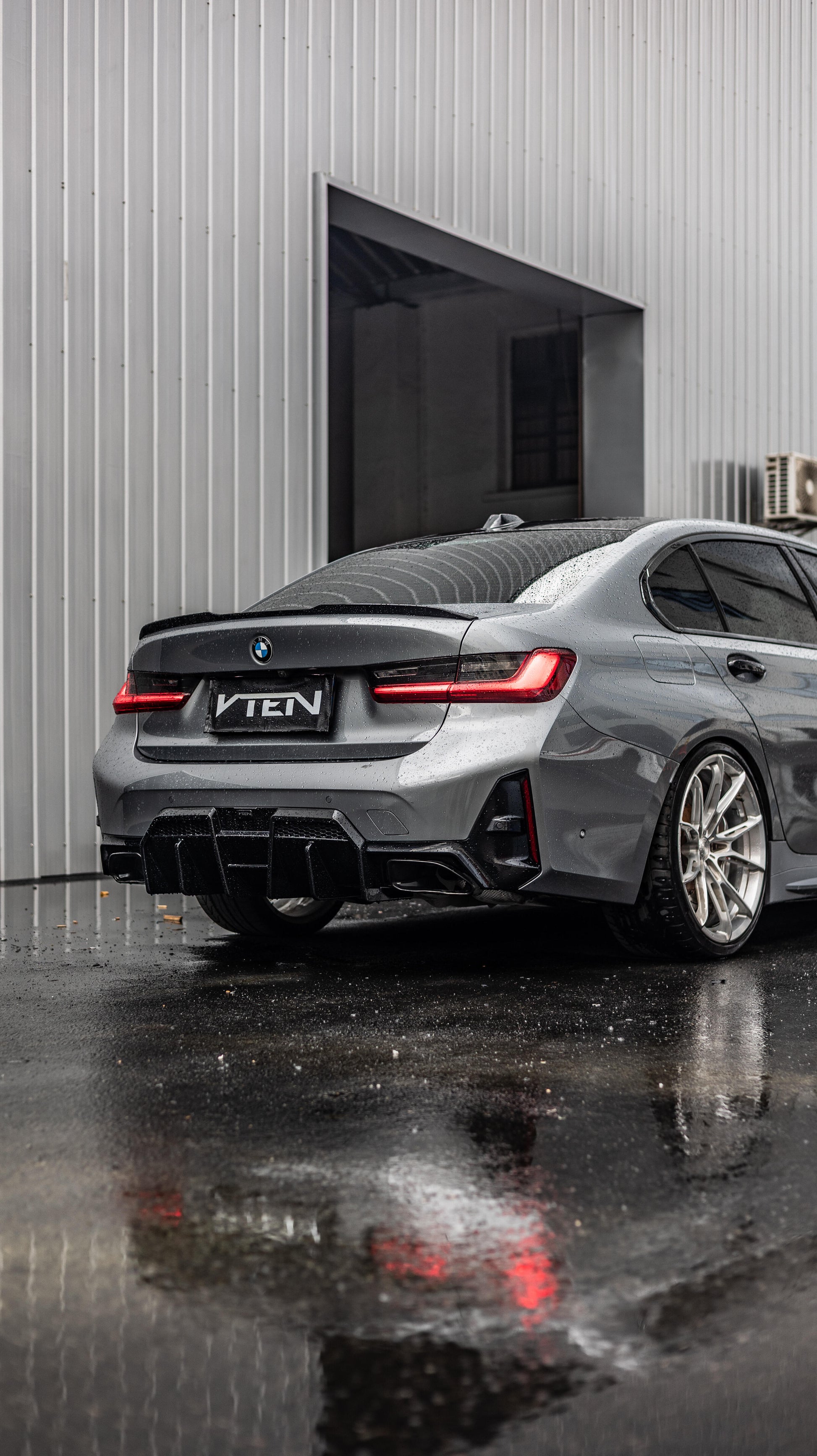 A dark gray BMW 3 Series G20 LCI sedan shown from the rear three-quarter angle, parked on wet pavement in front of a gray corrugated metal building. The car features a black rear diffuser, a custom black VTEN badge on the trunk, and multi-spoke alloy wheels.