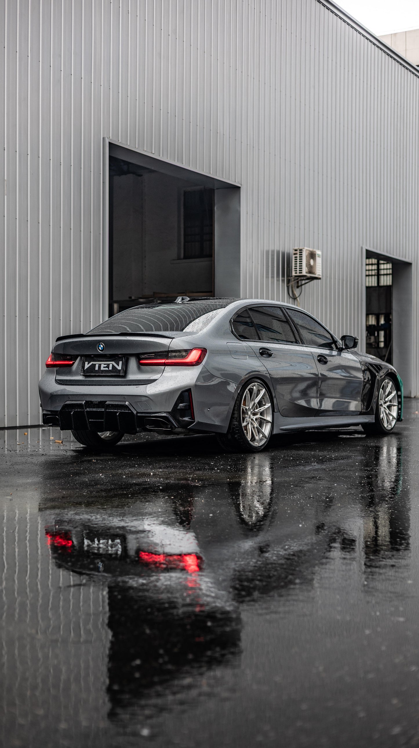 A dark gray BMW 3 Series sedan with a custom black VTEN wrap on the rear and sides, parked on wet asphalt in front of a gray metal building. The car is shown from a rear three-quarter angle, with its reflection clearly visible on the wet ground.