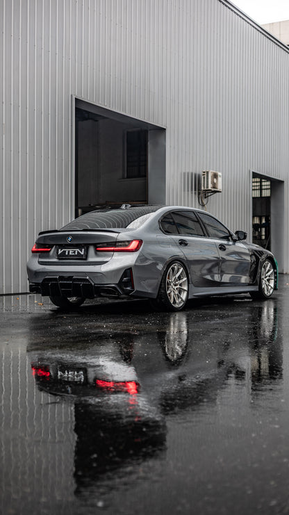 A dark gray BMW 3 Series sedan with a custom black VTEN wrap on the rear and sides, parked on wet asphalt in front of a gray metal building. The car is shown from a rear three-quarter angle, with its reflection clearly visible on the wet ground.