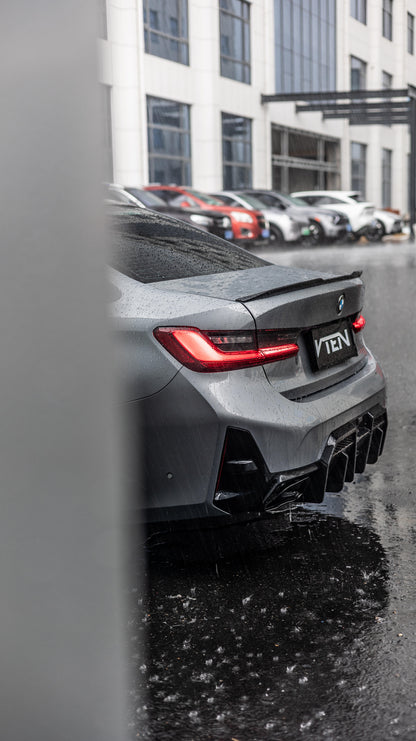 Rear three-quarter view of a dark gray BMW 3 Series G20 LCI with a custom black diffuser and a 'VTEN' license plate, parked on a wet street in front of a modern building.