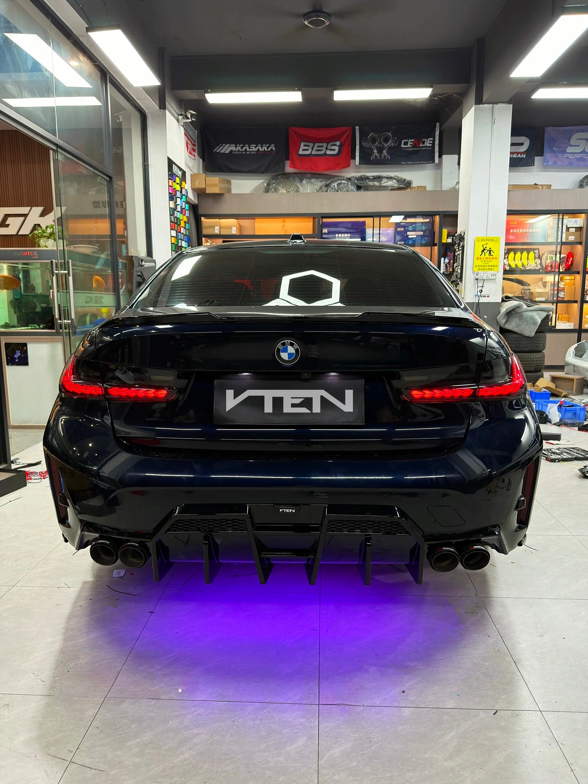 Rear view of a black BMW  BMW 3 Series G20 LCI with a large rear diffuser and custom license plate frame reading VTEN, parked inside a car workshop with purple underglow lighting.