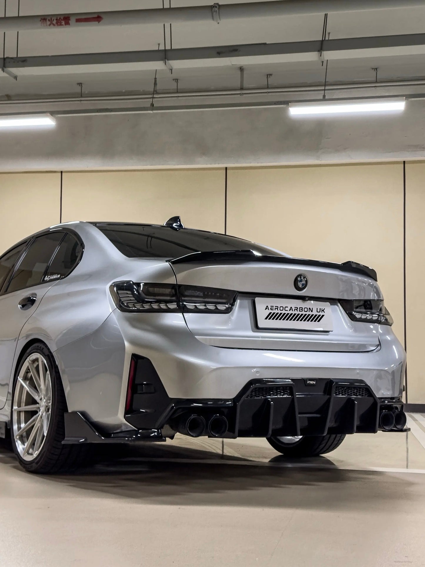 A silver BMW 3 Series G20 LCI sedan shown from the rear in an indoor parking structure, fitted with a black carbon fiber rear diffuser, rear spoiler, and side skirts by AEROCARBONUK.